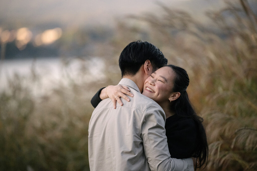 She smiles over his shoulder in a close embrace, autumn grass and the pale water of Lake Kawaguchi softly blurred behind them.