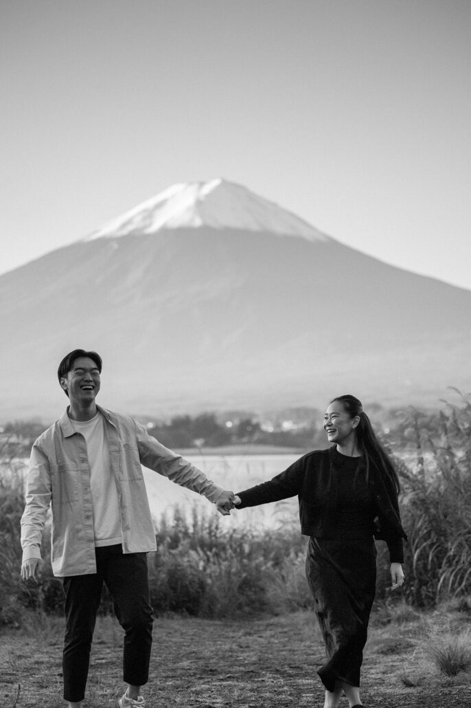 In black and white, the newly engaged couple laughs as they walk hand in hand by Lake Kawaguchi with Mt Fuji's snowy peak behind.