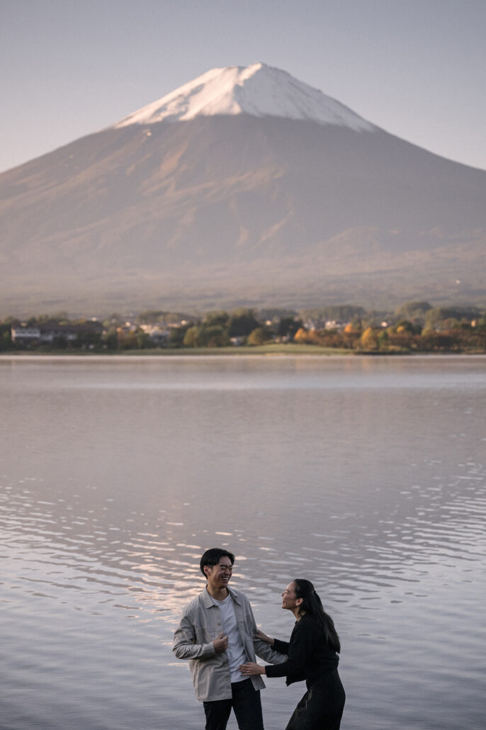 The couple laughs together at the edge of Lake Kawaguchi, Mt Fuji rising above the far shore in a tall autumn engagement frame.