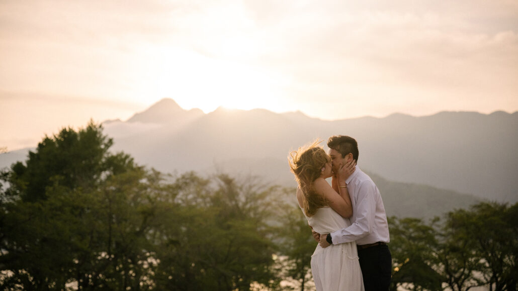 The couple kisses as the sun sets behind the mountains near Kawaguchiko, green spring trees below the hazy, glowing ridge.