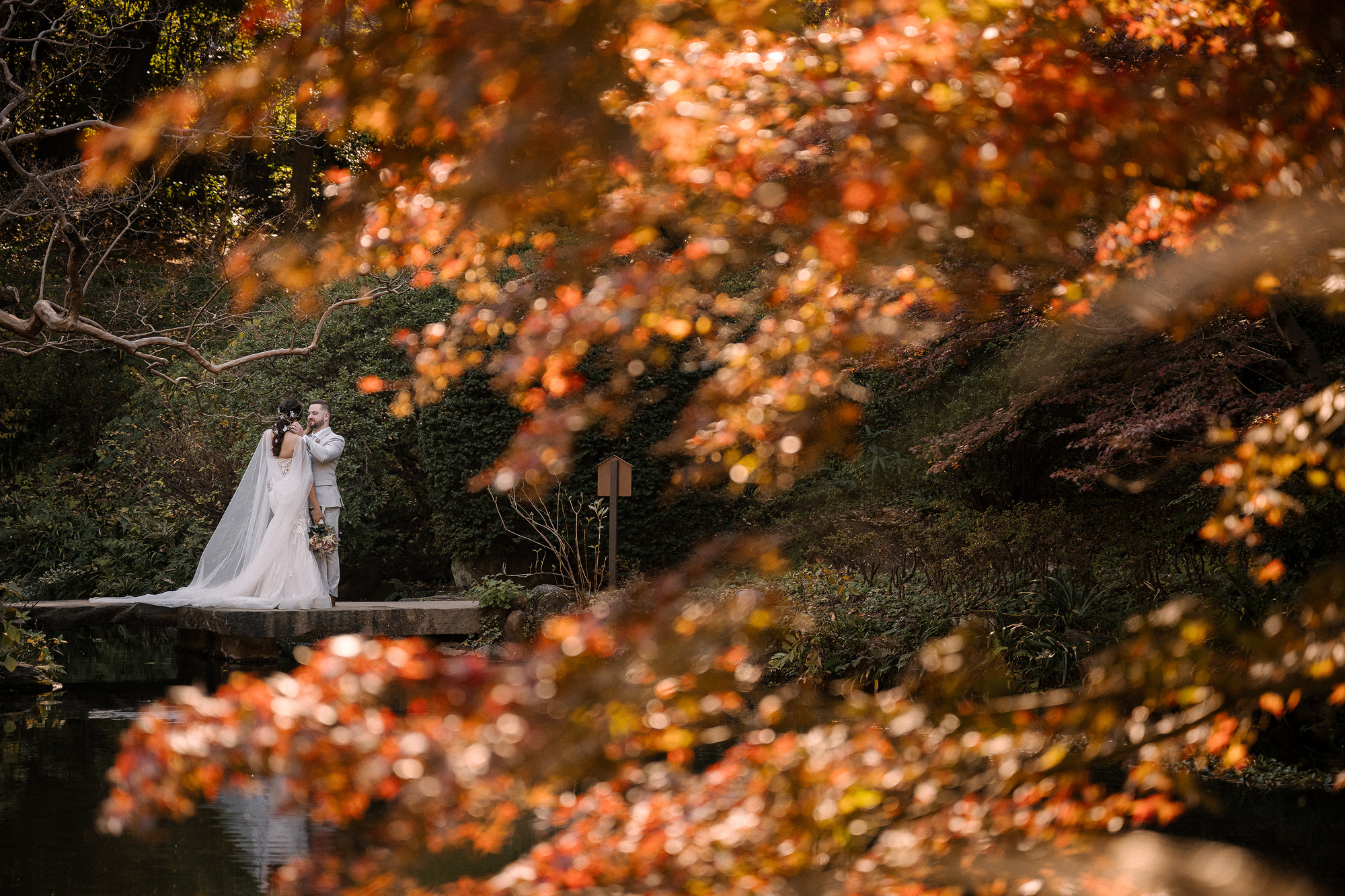 The couple stand on a stone path by the pond, framed by red and orange maples, after their symbolic ceremony in a Tokyo garden.