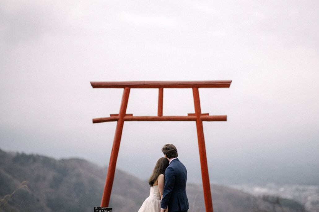 The couple stand together beneath the red gate of Tenku no Torii, hills and low cloud beyond, during their Mt Fuji elopement.