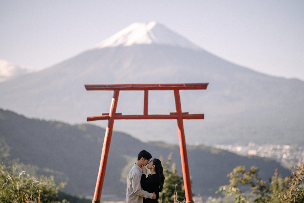 The couple kisses between the red posts of Tenku no Torii, Mt Fuji soft behind them and the valley town far below.
