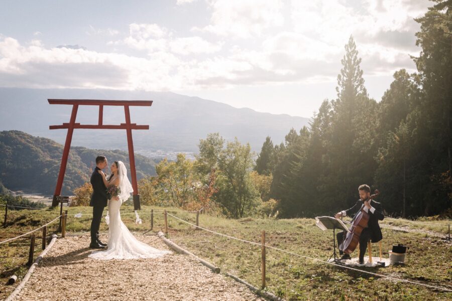 The bride and groom embrace under the red torii as a cellist plays beside them, the peak of Mt Fuji above the cloud at their Tenku no Torii wedding.