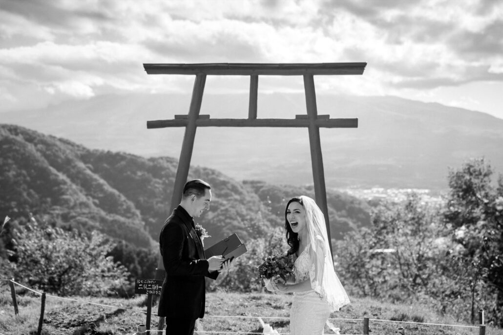 In black and white, the bride laughs as the groom reads his vows from a small book at their Tenku no Torii wedding, trees and hills behind them.