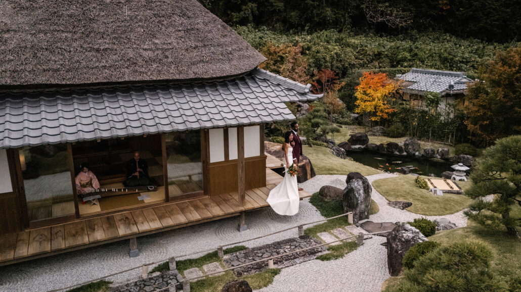 Seen from above, the bride's veil swirls beside a thatched villa where a musician plays inside, the garden turning red and gold.