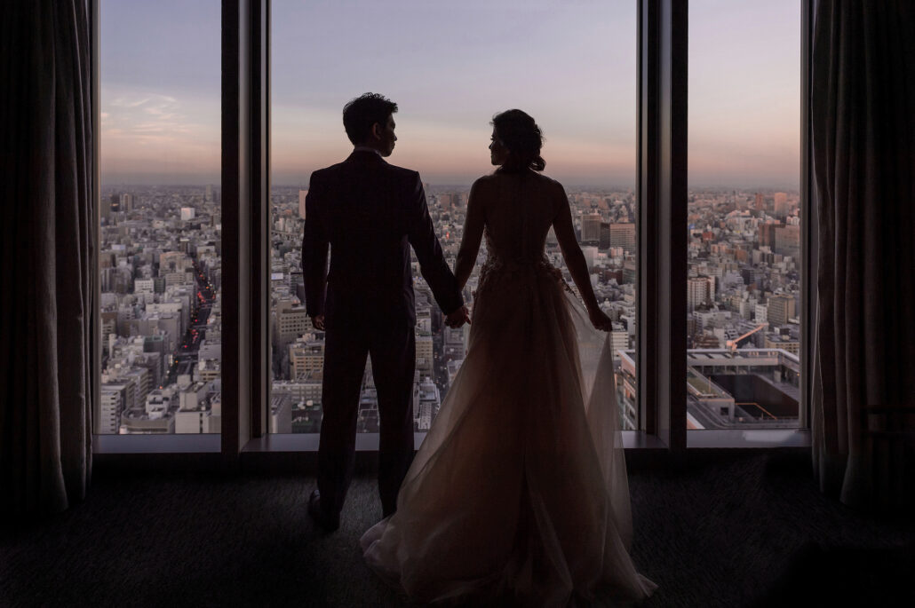 The bride and groom stand hand in hand at a tall window high above Tokyo as the light fades.