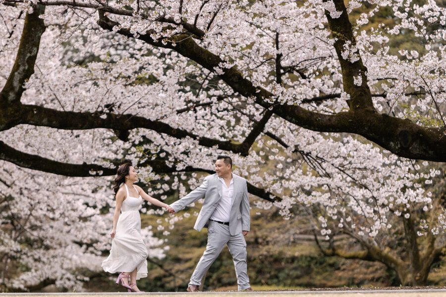 The couple walk hand in hand beneath a wide arch of cherry blossom branches in a Tokyo garden, a classic moment for Tokyo engagement photos.