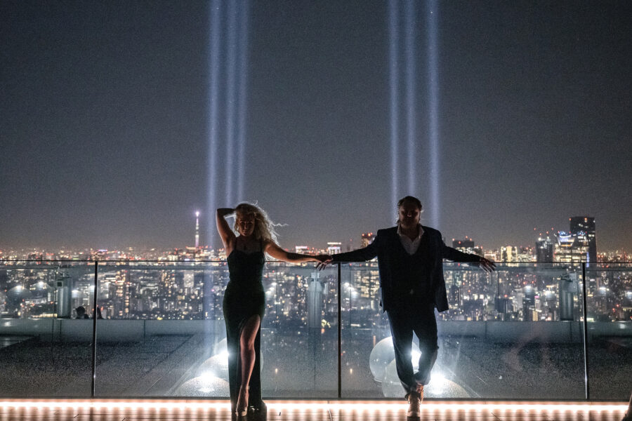 An engaged couple on the glass edge of the Shibuya Sky rooftop at night, searchlight beams rising over the Tokyo skyline behind them.