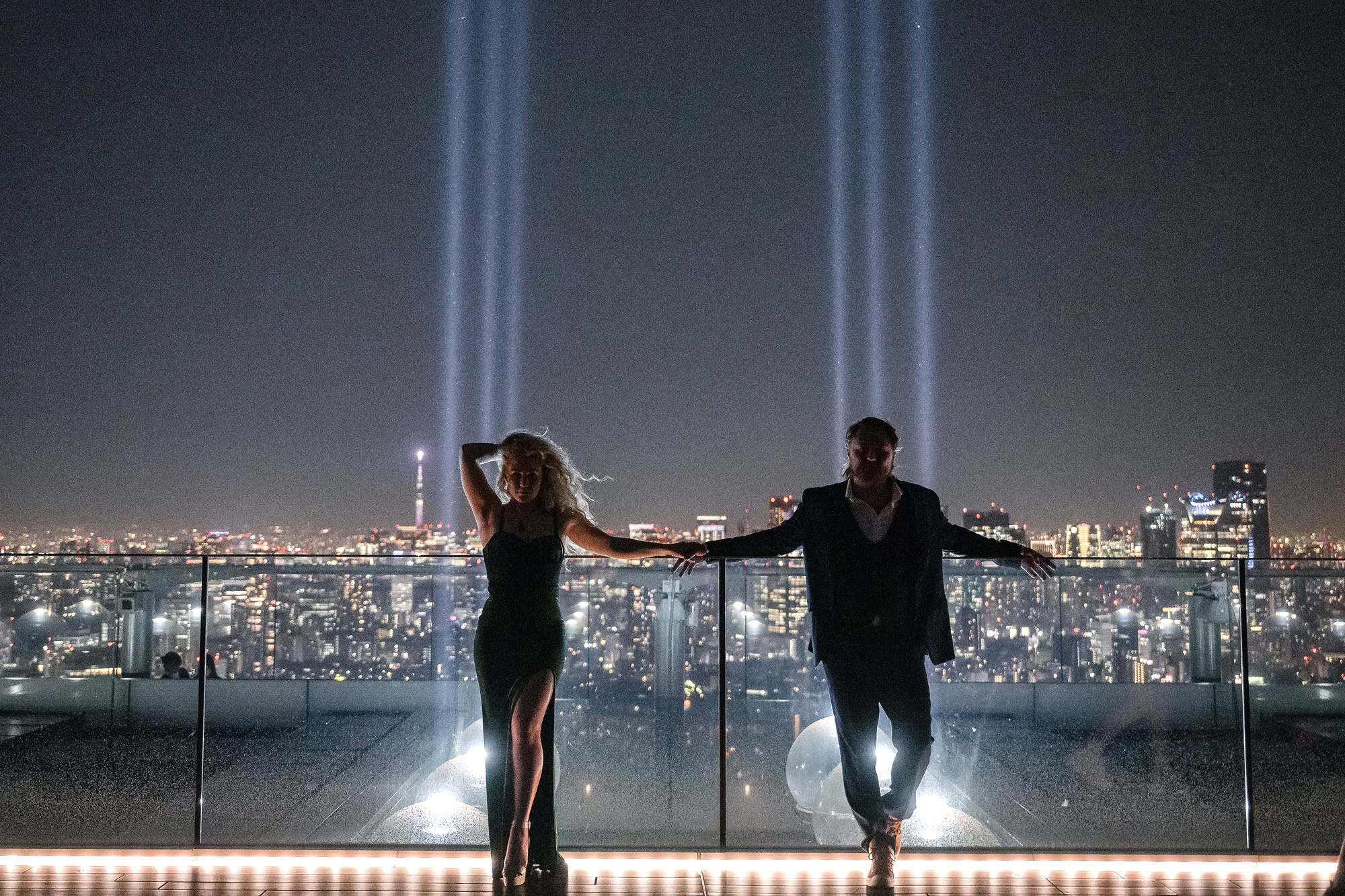 An engaged couple on the glass edge of the Shibuya Sky rooftop at night, searchlight beams rising over the Tokyo skyline behind them.