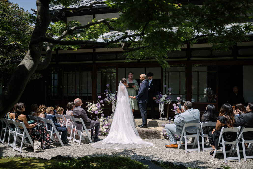 Guests sit in a garden shaded by trees as the bride and groom marry in front of a traditional Japanese house at a garden wedding in Tokyo.