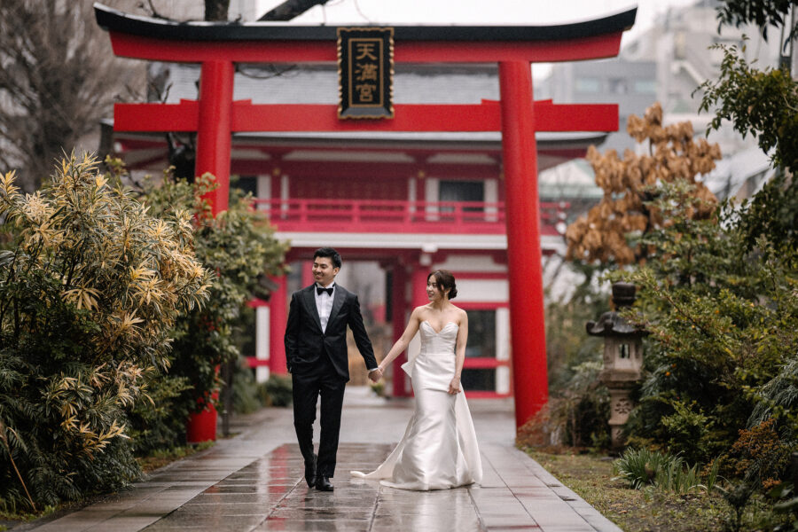 Hand in hand, the bride and groom walk out from beneath the vermilion torii of a shrine in Tokyo on a wet day.