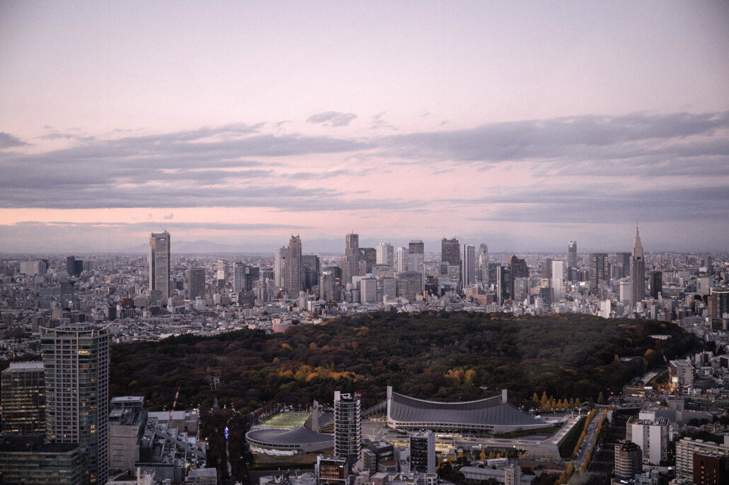 Tokyo at dusk from Shibuya Sky, a stadium and a green forest in front of the city lights.