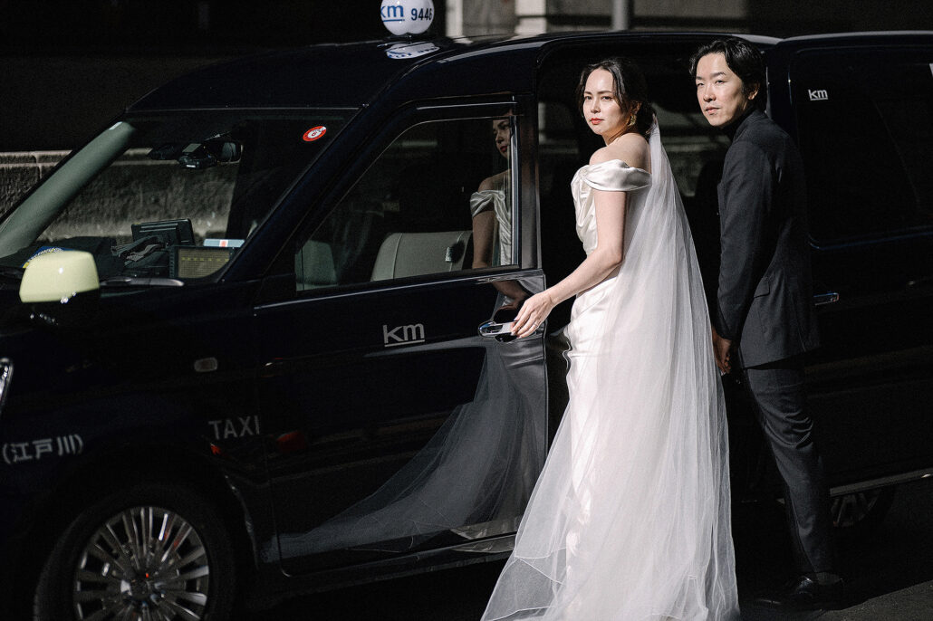 The bride and groom step out of a black Tokyo taxi at night, her long veil spilling onto the street.