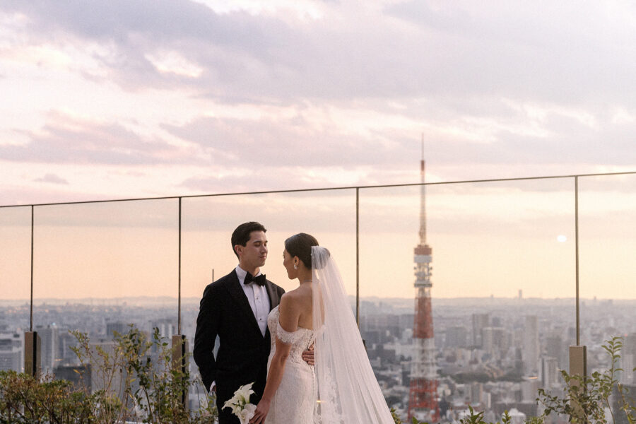The bride and groom on the rooftop at sunset, her long veil across the terrace, Tokyo Tower rising behind at a Toranomon Hills wedding.
