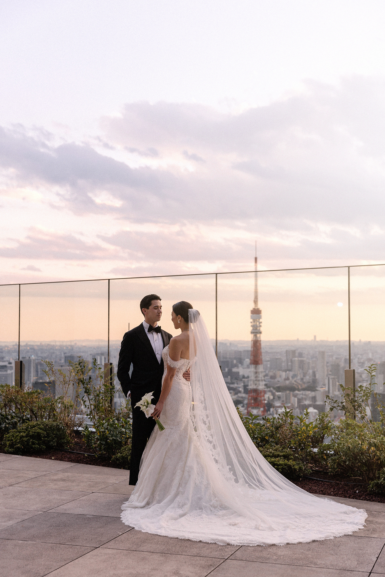 The bride and groom on the rooftop at sunset, her long veil across the terrace, Tokyo Tower rising behind at a Toranomon Hills wedding.