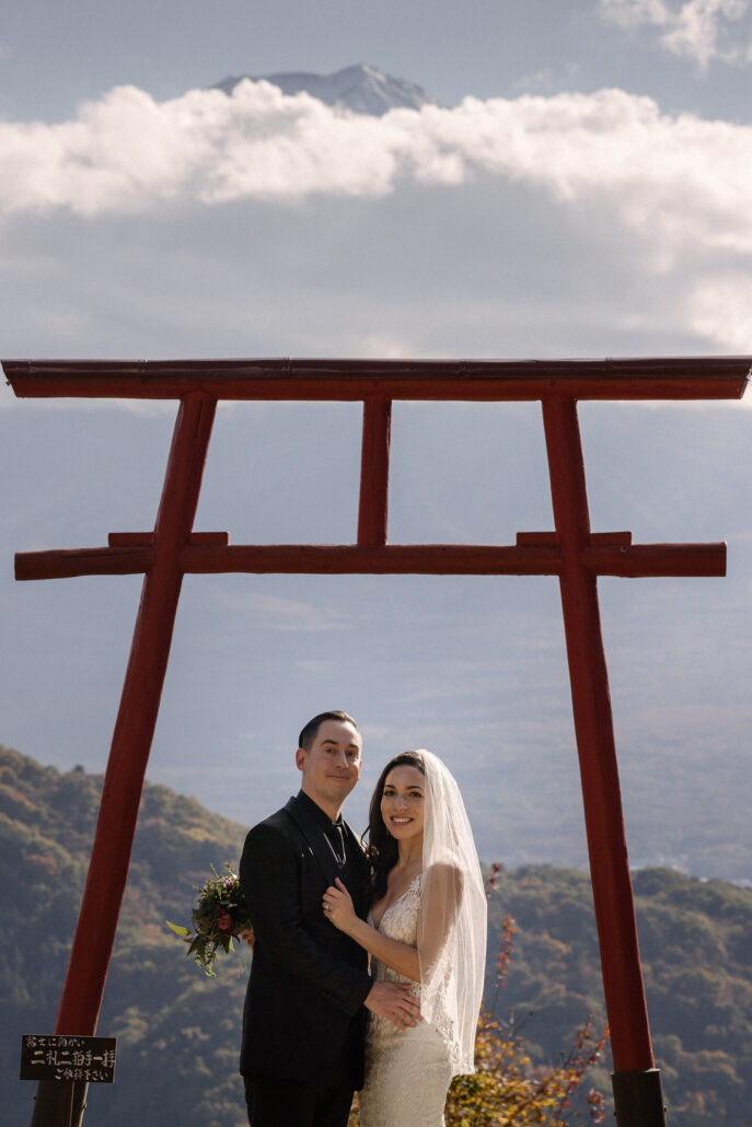 The couple stand beneath the red gate at their Tenku no Torii wedding as the summit of Mt Fuji appears above a band of cloud.