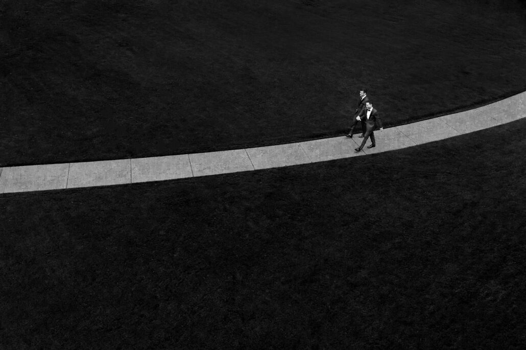 Two grooms walk hand in hand along a curving path across a dark lawn, small in the frame, in black and white.