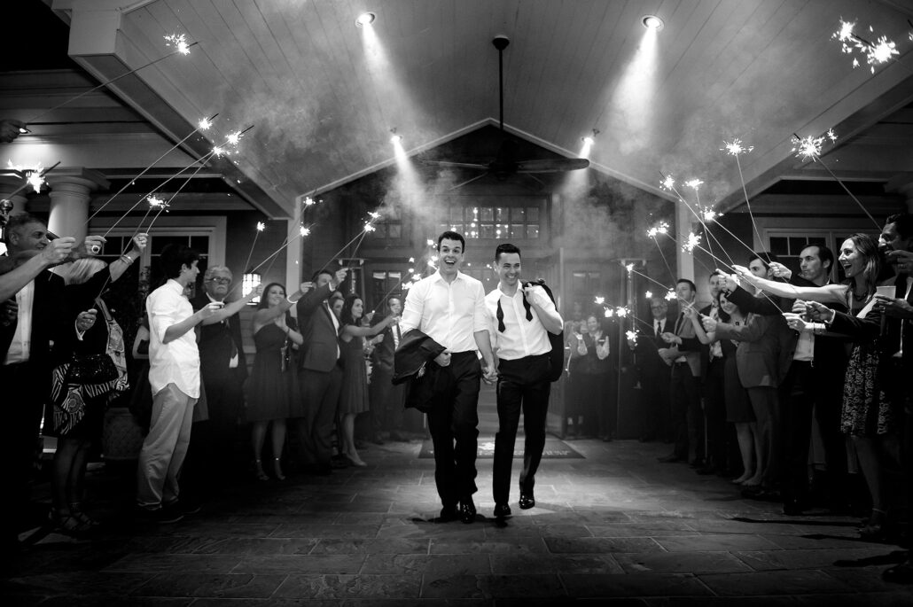 In black and white, two grooms walk out through a tunnel of sparklers held high by their guests.