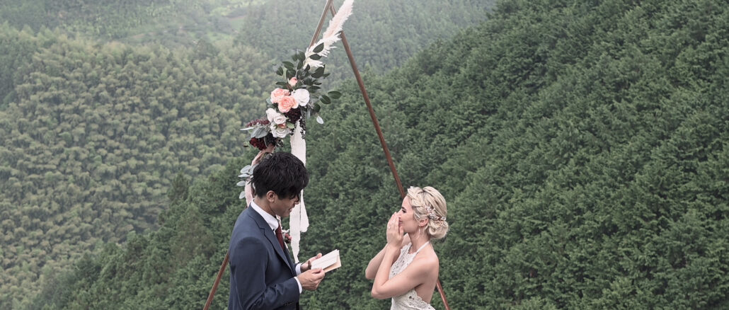 The groom reads his vows from a card as the bride covers her face with her hands, at a flower arch before a forested mountainside.
