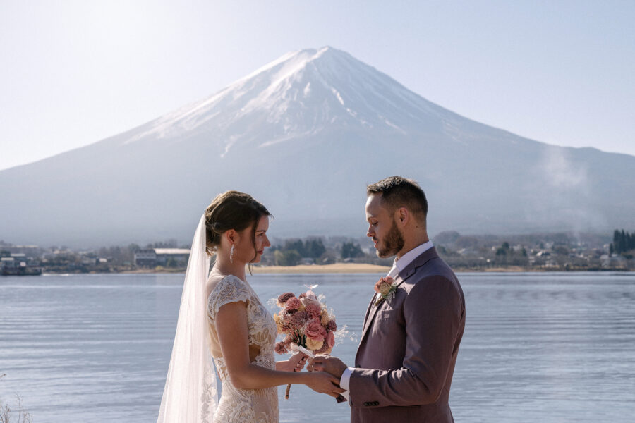 The bride holds her pink bouquet as she faces the groom by Lake Kawaguchi, Mt Fuji white and close behind, a December elopement in Japan.