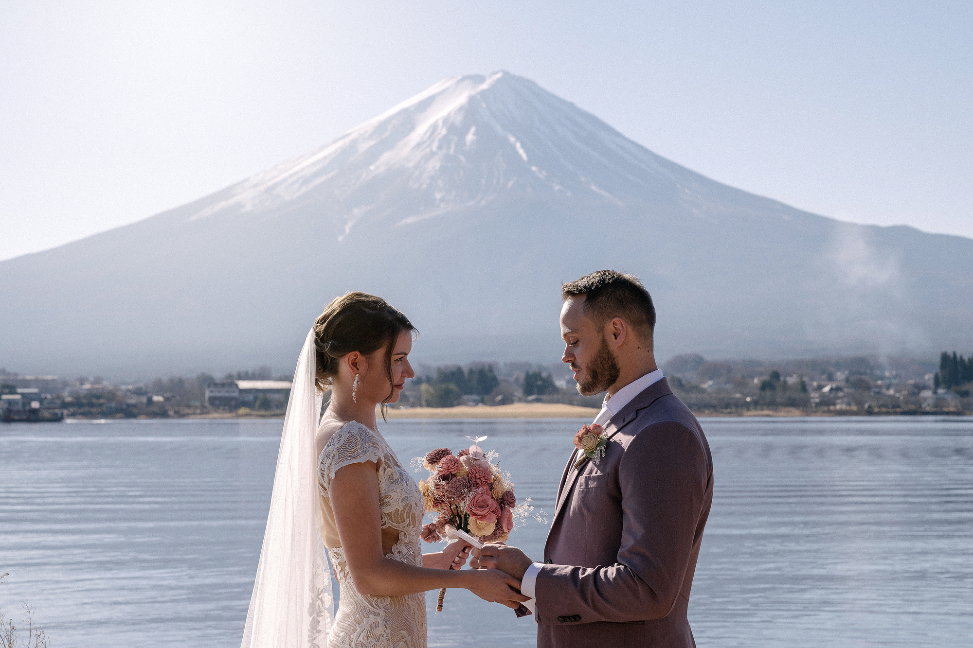 The bride holds her pink bouquet as she faces the groom by Lake Kawaguchi, Mt Fuji white and close behind, a December elopement in Japan.