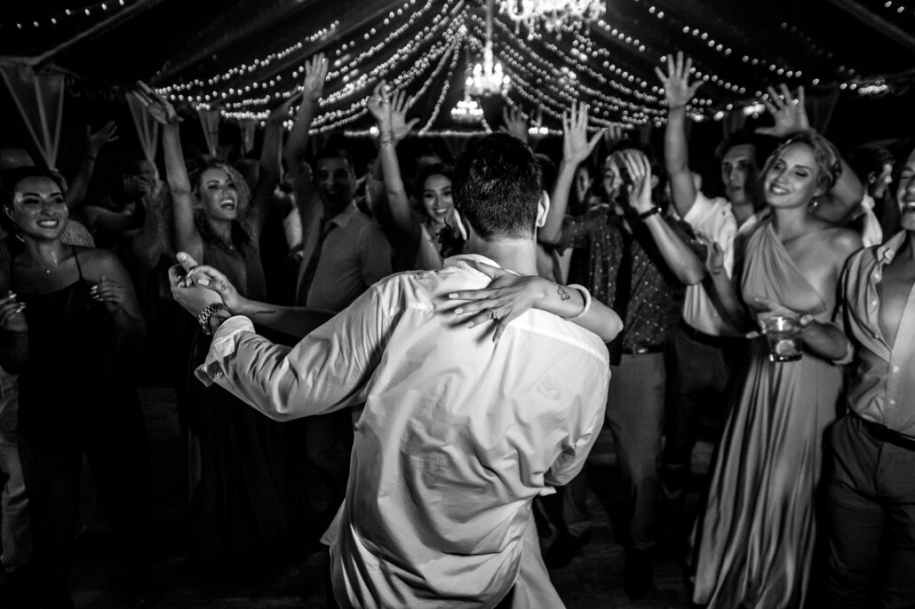 In black and white, the couple hold each other on the dance floor as guests throw their hands up beneath a canopy of string lights.