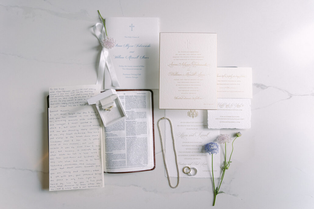 The wedding details laid out on white marble: the invitation suite, a handwritten letter of vows, the rings, a pearl necklace and sprigs of scabiosa.