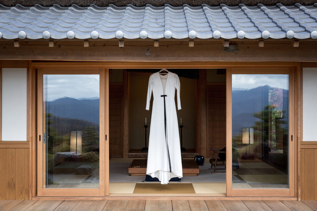The wedding dress hangs in the open doorway of a traditional Japanese house, with mountains through the windows on either side.