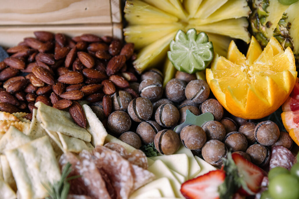 Close up on a wedding grazing table: almonds, macadamias, pineapple, a lime and an orange cut into flowers, crackers, cheese and strawberries.