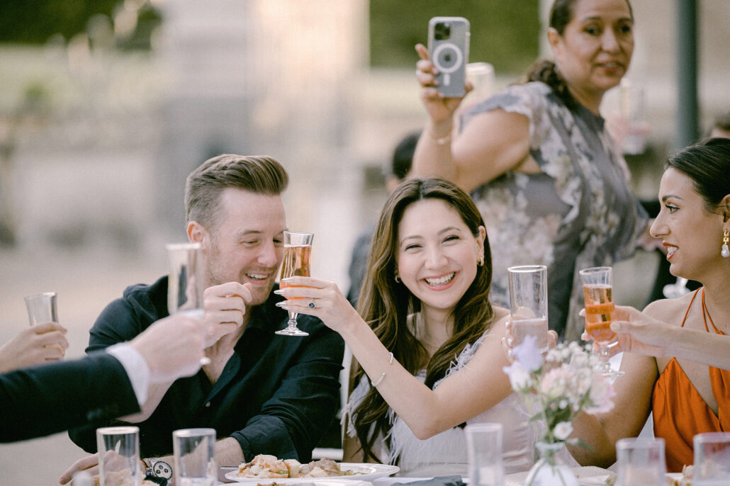 Guests raise their glasses and laugh across a long table at a wedding dinner in France.