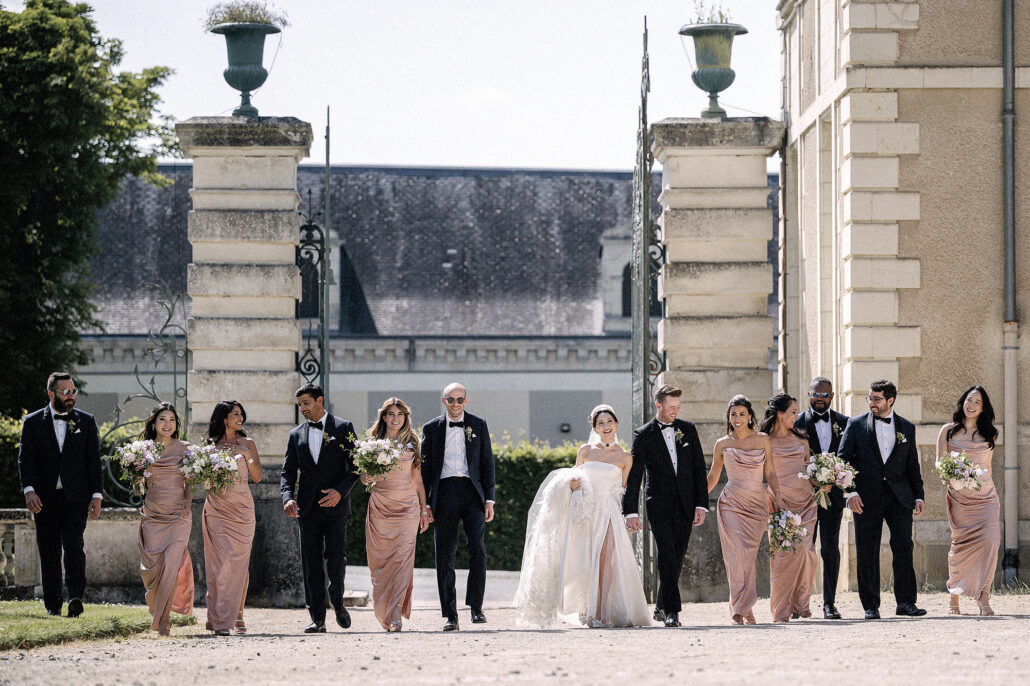 The bride and groom walk through the stone gates of a French chateau with their wedding party in dusky pink and black tie.