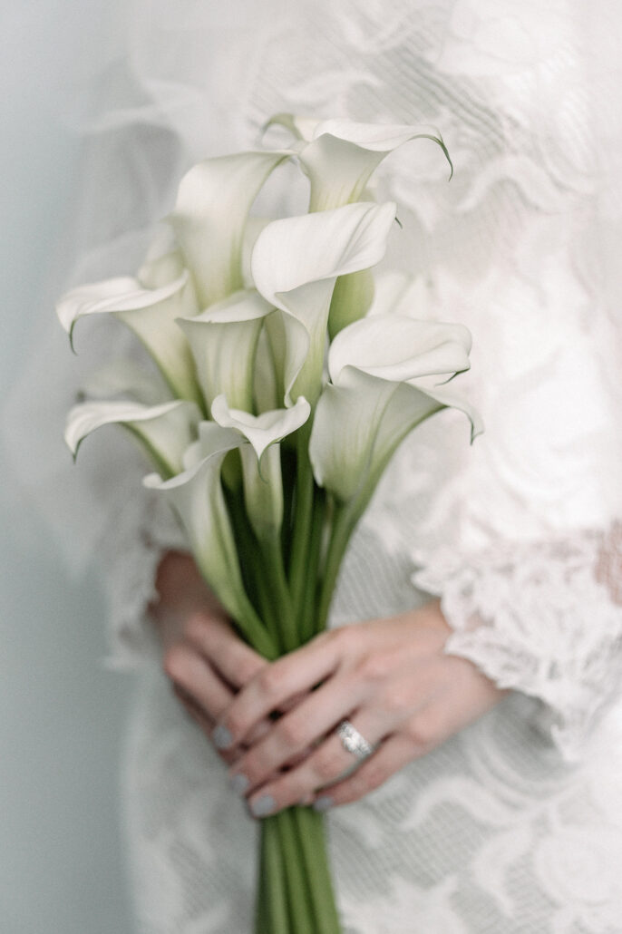 The bride holds a bouquet of long-stemmed white calla lilies against the lace of her sleeves.