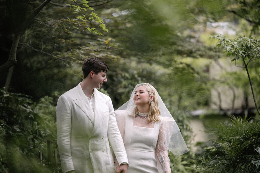 Leaves blow across the frame as the couple stand together in the garden, wind moving through the trees on an August elopement day.