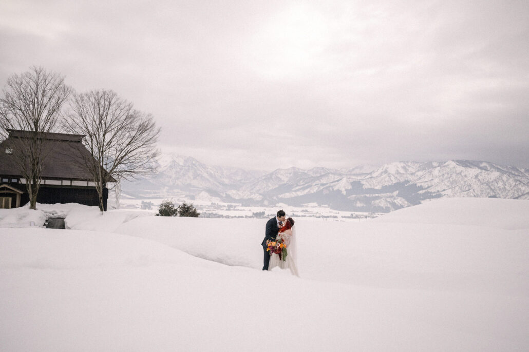 The bride and groom kiss in a wide field of snow beside a traditional Japanese house, mountains behind them, on a winter elopement in Japan.